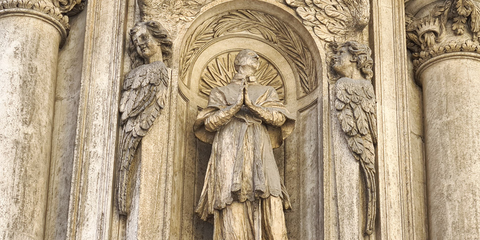 Saint Charles Borromeo stands in prayer framed by sculptural angels